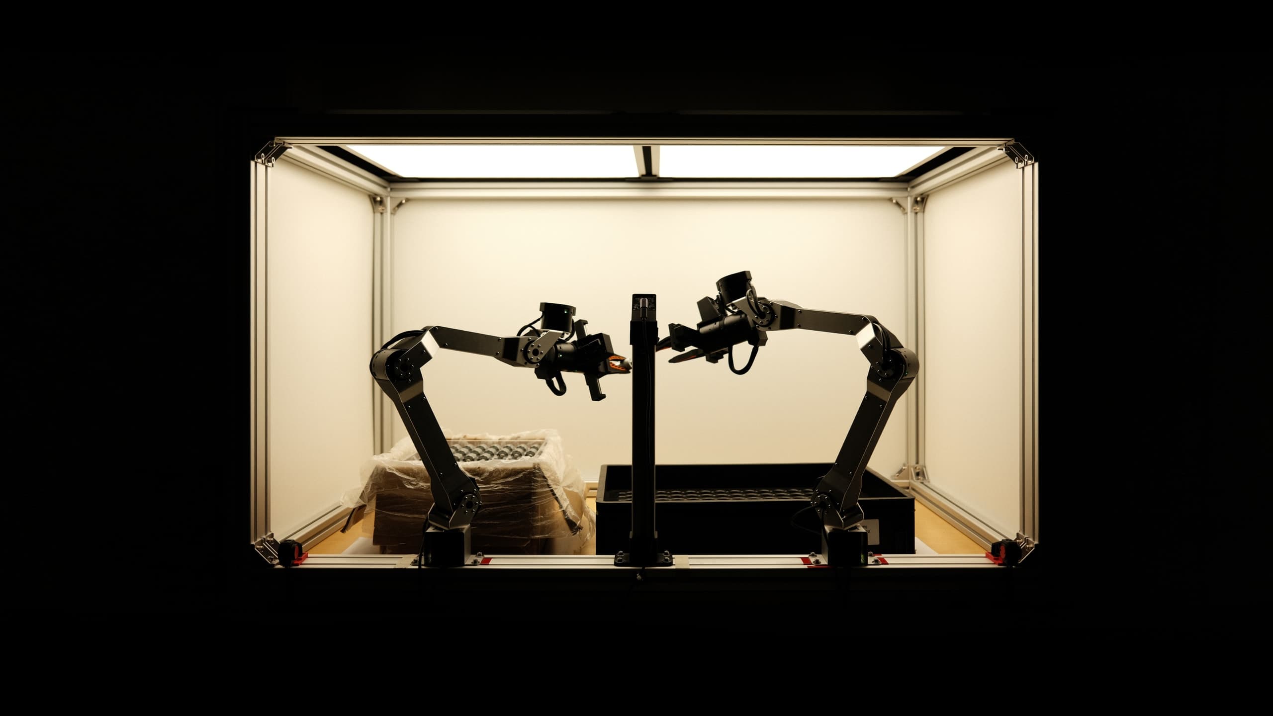 Two robot arms facing each other inside a lit photo box, one holding an actuator mid-handover, with the shipping carton on the left and the parts tray on the right.